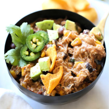 creamy chili made with chicken, taco seasoning, corn, beans, tomatoes, cream cheese in a bowl topped with avocado, cilantro and chips