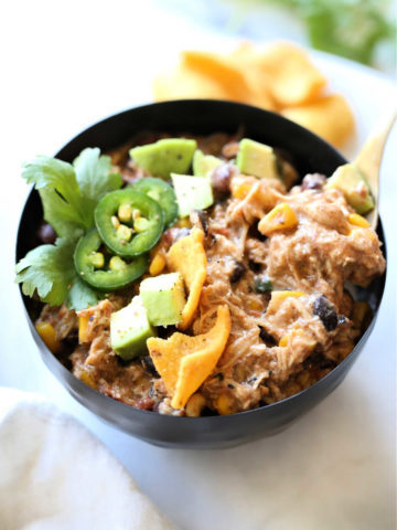 creamy chili made with chicken, taco seasoning, corn, beans, tomatoes, cream cheese in a bowl topped with avocado, cilantro and chips