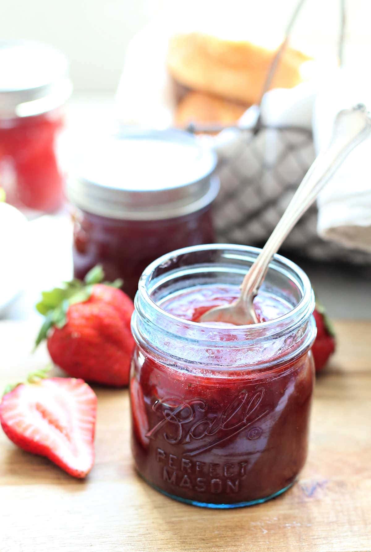homemade strawberry jam in a jar with a spoon and fresh sliced strawberries next to it