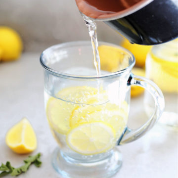 saucepan pouring hot water over lemon juice and lemon citrus slices in a clear mug