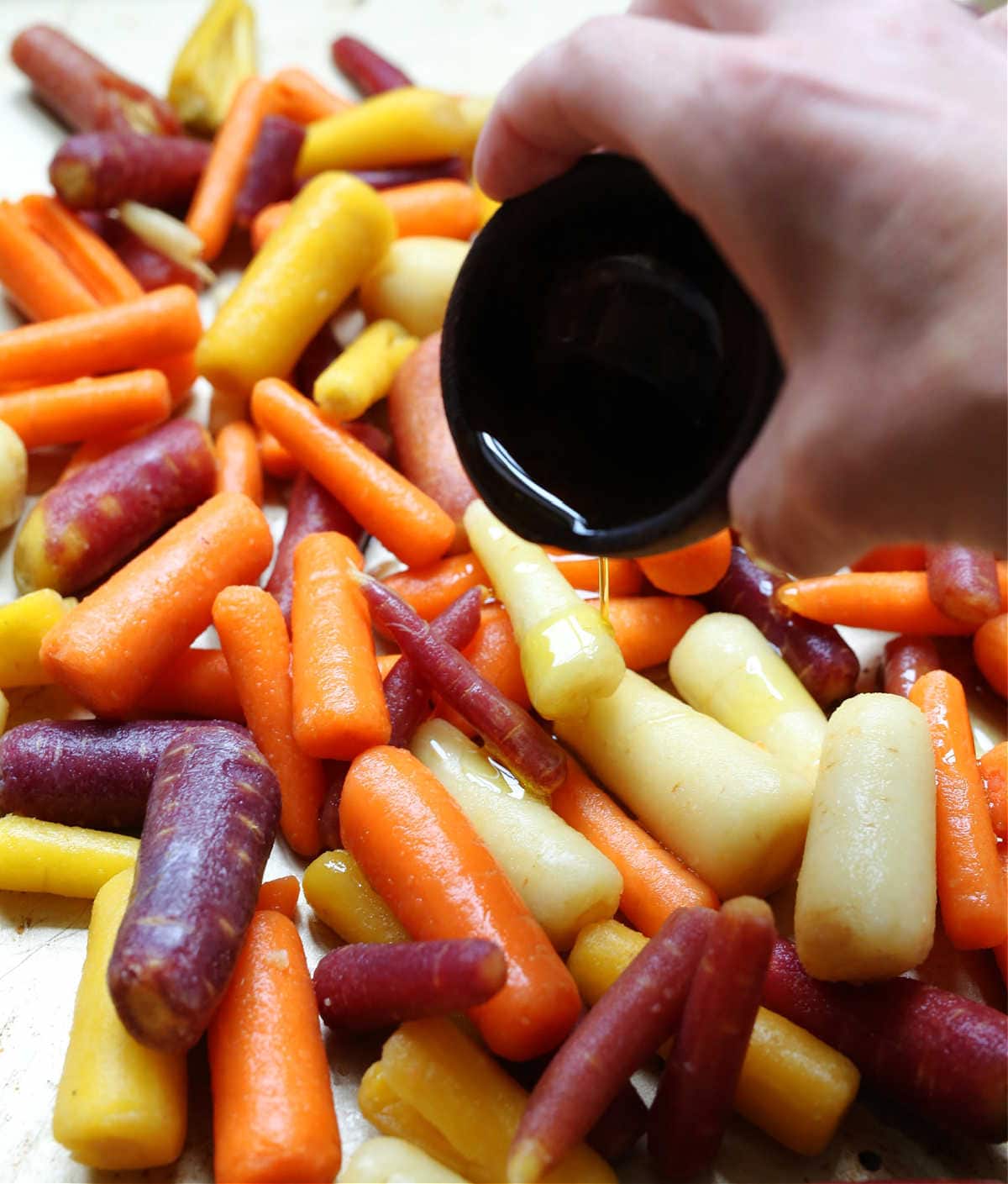 multi colored carrots being topped with olive oil in preparation to be roasted