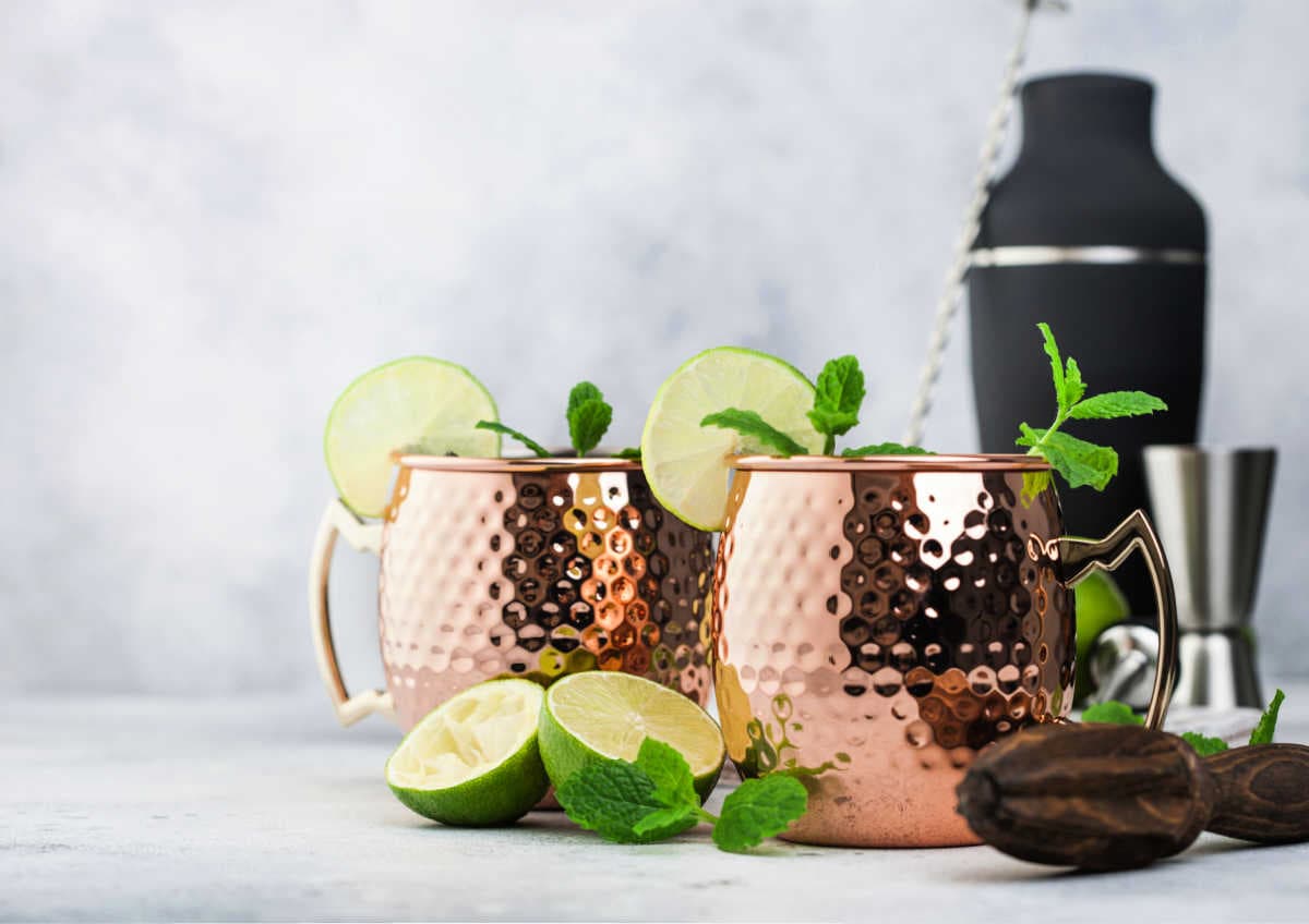 two mugs filled with a moscow mule recipe and limes, cocktail mixer, and fresh mint on a table