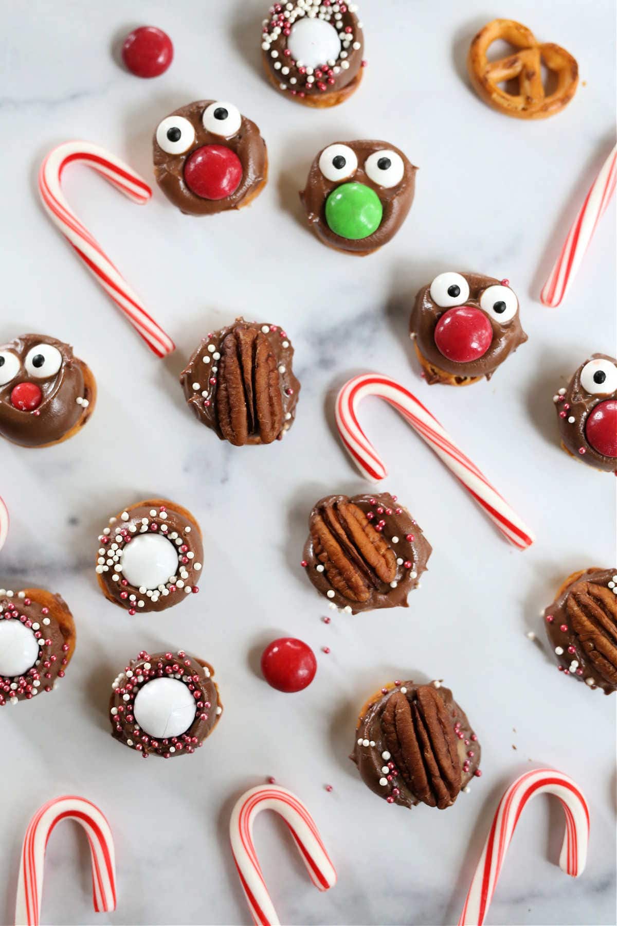 rolo candy and chocolate caramel candies topped on pretzels with pecans and candy decorations on a marble surface surrounded by candy canes