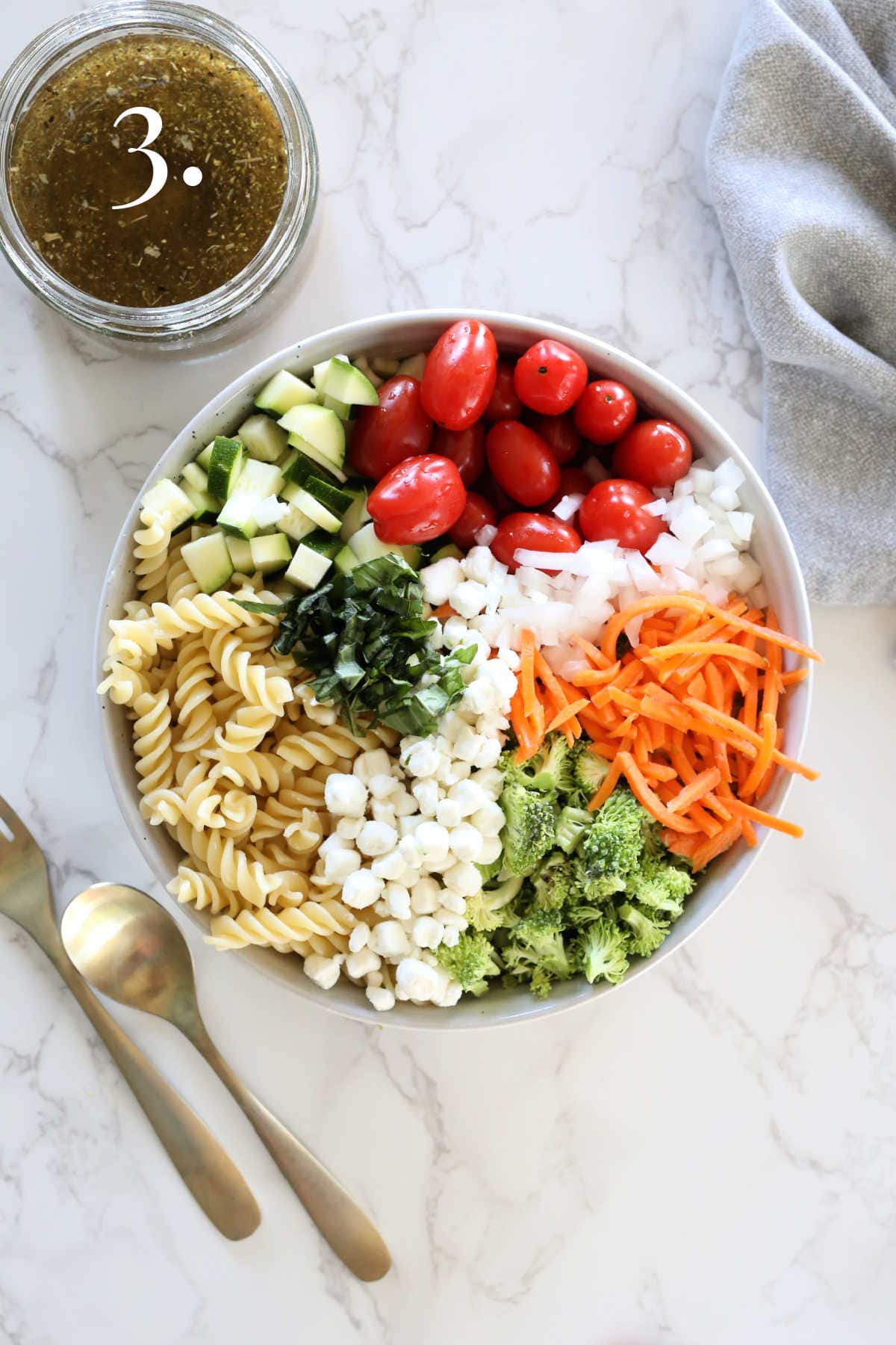 Pasta salad ingredients in a salad bowl with silverware and Italian dressing.
