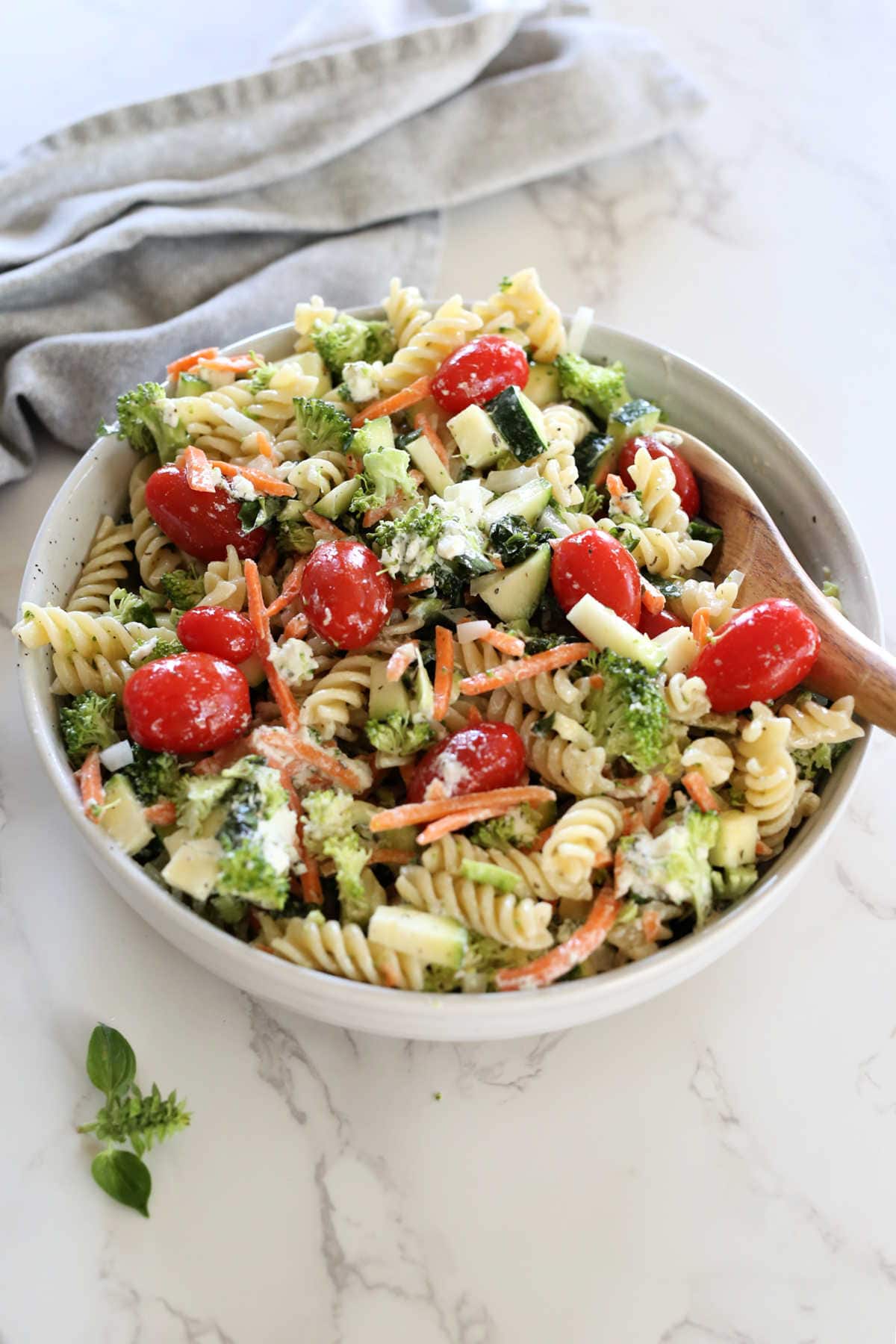 Pasta salad made with gluten-free noodles, cucumbers, zucchini, tomatoes, goat cheese, Italian dressing in a bowl with a serving spoon.