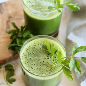 Parsley juice with celery and cucumber in two glasses garnished with flat parsley leaves on stem.