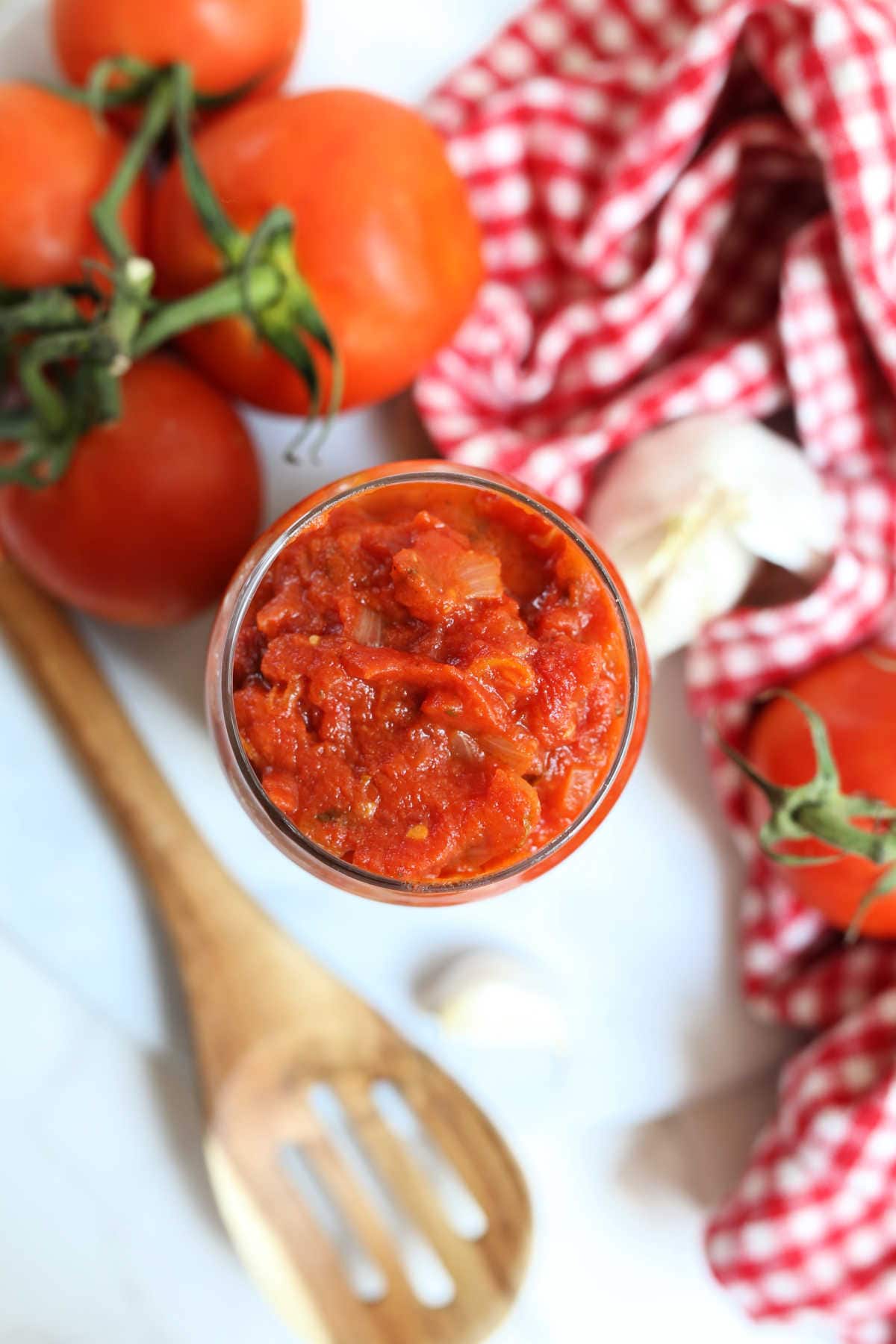 Homemade marinara sauce recipe in a glass jar with tomatoes towel and spoon.