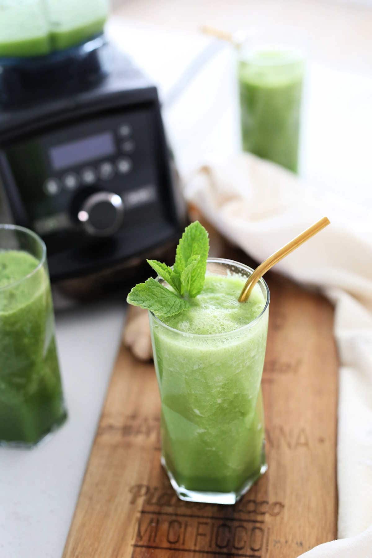 Green juiced recipes in glasses on a cutting board and counter next a Vitamix blender.