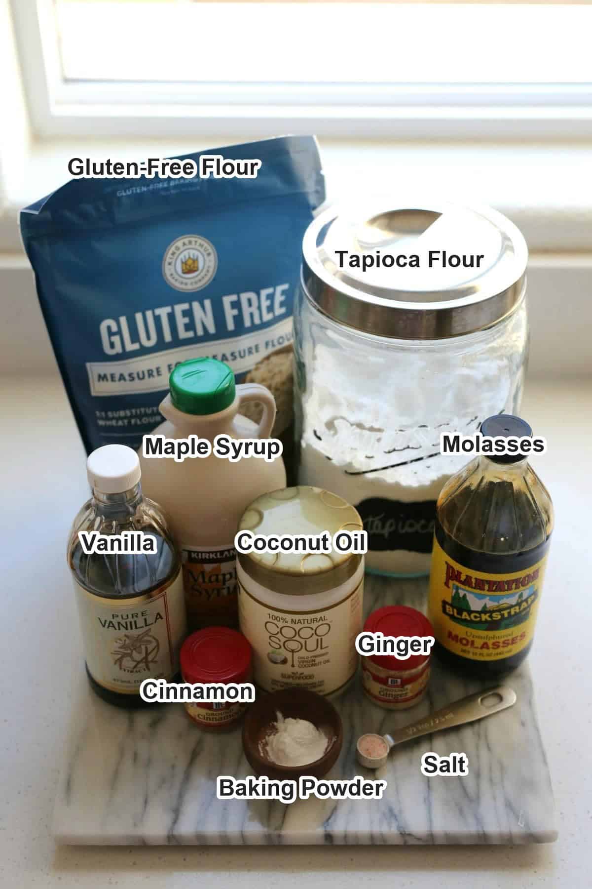 Labeled ingredients for a gluten-free gingerbread house: gluten-free flour, tapioca flour, maple syrup, molasses, vanilla, coconut oil, cinnamon, ginger, baking powder and salt.