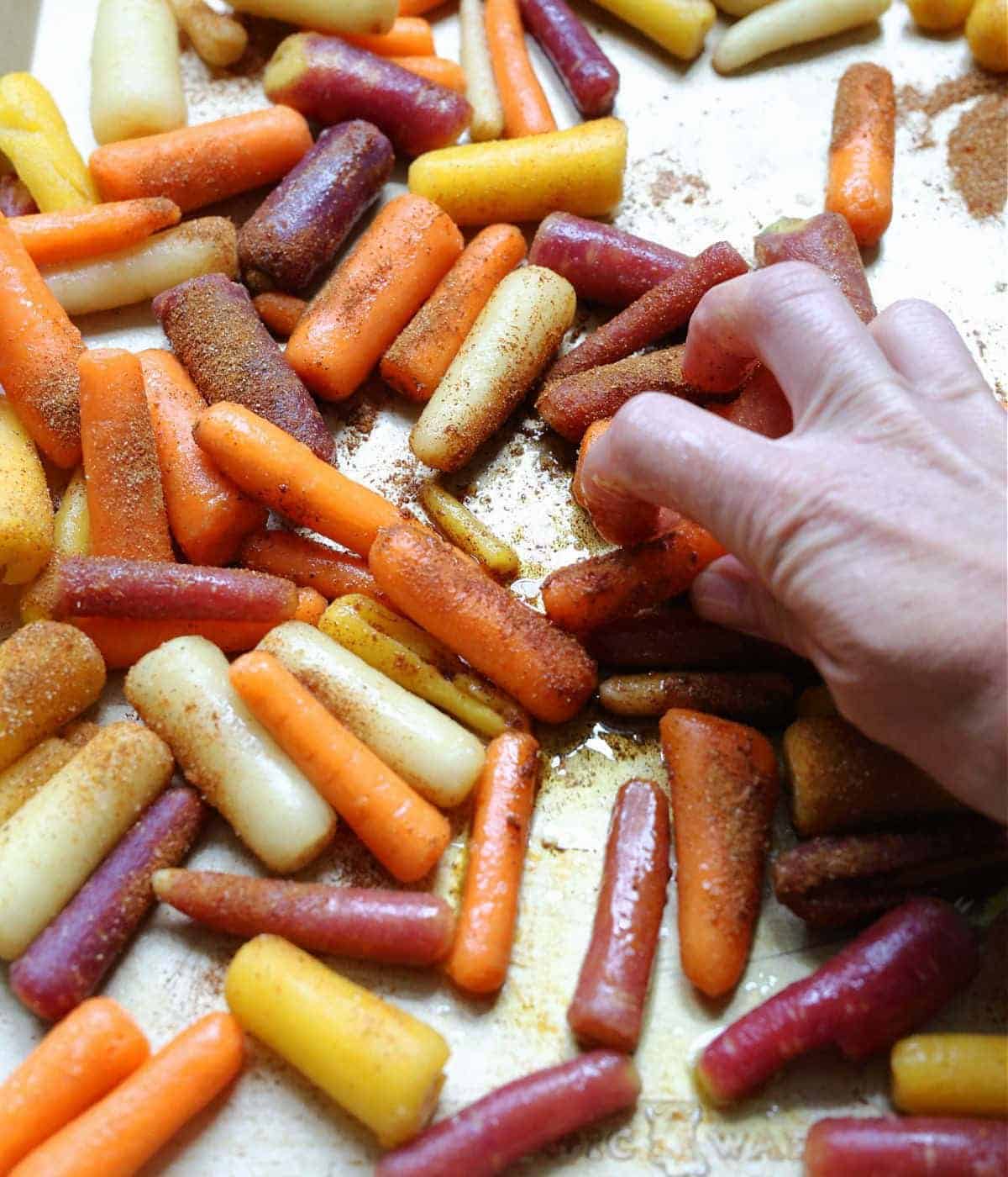 Hand tossing rainbow carrots in oil and seasonings on the baking sheet.